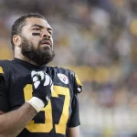 Cam Heyward, über 30 und mit der Nummer 97 im schwarz-gelben Steelers-Trikot, steht mit der Hand auf der Brust auf dem Spielfeld und blickt nach oben, während die Menschenmenge im Stadionhintergrund verschwimmt. Diese Beschreibung wurde mit der FootballR KI automatisch generiert.