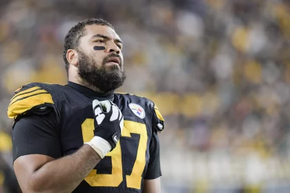 Cam Heyward, über 30 und mit der Nummer 97 im schwarz-gelben Steelers-Trikot, steht mit der Hand auf der Brust auf dem Spielfeld und blickt nach oben, während die Menschenmenge im Stadionhintergrund verschwimmt. Diese Beschreibung wurde mit der FootballR KI automatisch generiert.
