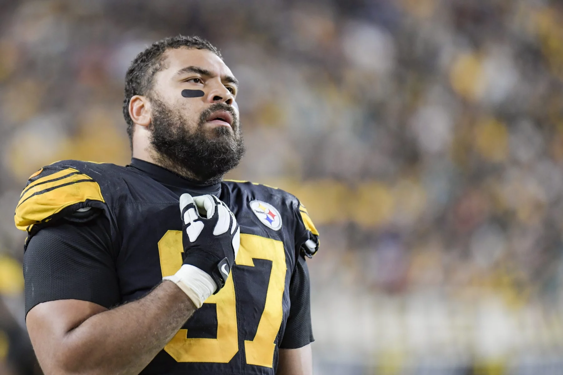 Cam Heyward, über 30 und mit der Nummer 97 im schwarz-gelben Steelers-Trikot, steht mit der Hand auf der Brust auf dem Spielfeld und blickt nach oben, während die Menschenmenge im Stadionhintergrund verschwimmt. Diese Beschreibung wurde mit der FootballR KI automatisch generiert.