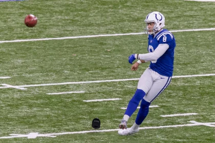 Kicker der Colts versucht einen Onside Kick - rechtes Bein ist ausgestreckt, während der Ball in der Luft über die weißen Yard-Linien und ein schwarzes Kicking-Tee auf dem Rasen fliegt. Diese Beschreibung wurde mit der FootballR KI automatisch generiert.