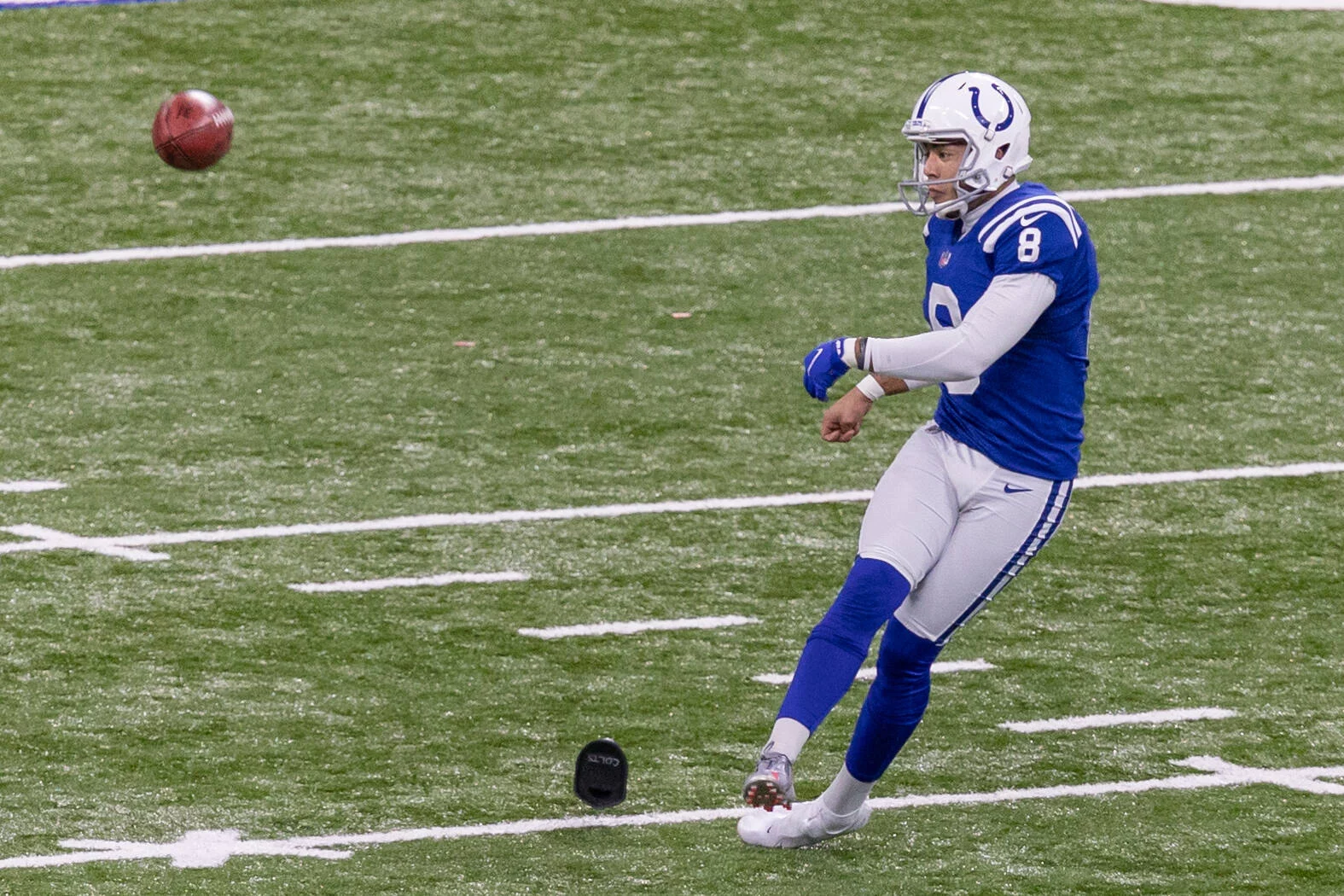 Kicker der Colts versucht einen Onside Kick - rechtes Bein ist ausgestreckt, während der Ball in der Luft über die weißen Yard-Linien und ein schwarzes Kicking-Tee auf dem Rasen fliegt. Diese Beschreibung wurde mit der FootballR KI automatisch generiert.
