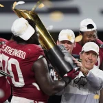 Kalen DeBoer will mit Alabama zurück an die Spitze. Eine Footballmannschaft in roten Trikots feiert den Gewinn der Nationalmeisterschaft. Die Spieler tragen weiße Hüte und halten einen großen goldenen Pokal in den Händen, während Konfetti um sie herum regnet. Ein Mann in weißer Jacke hilft, den Pokal zu halten, und lächelt zwischen den Spielern auf einer Bühne mit schwarz-weißem Hintergrund. Diese Beschreibung wurde mit der FootballR KI automatisch generiert.