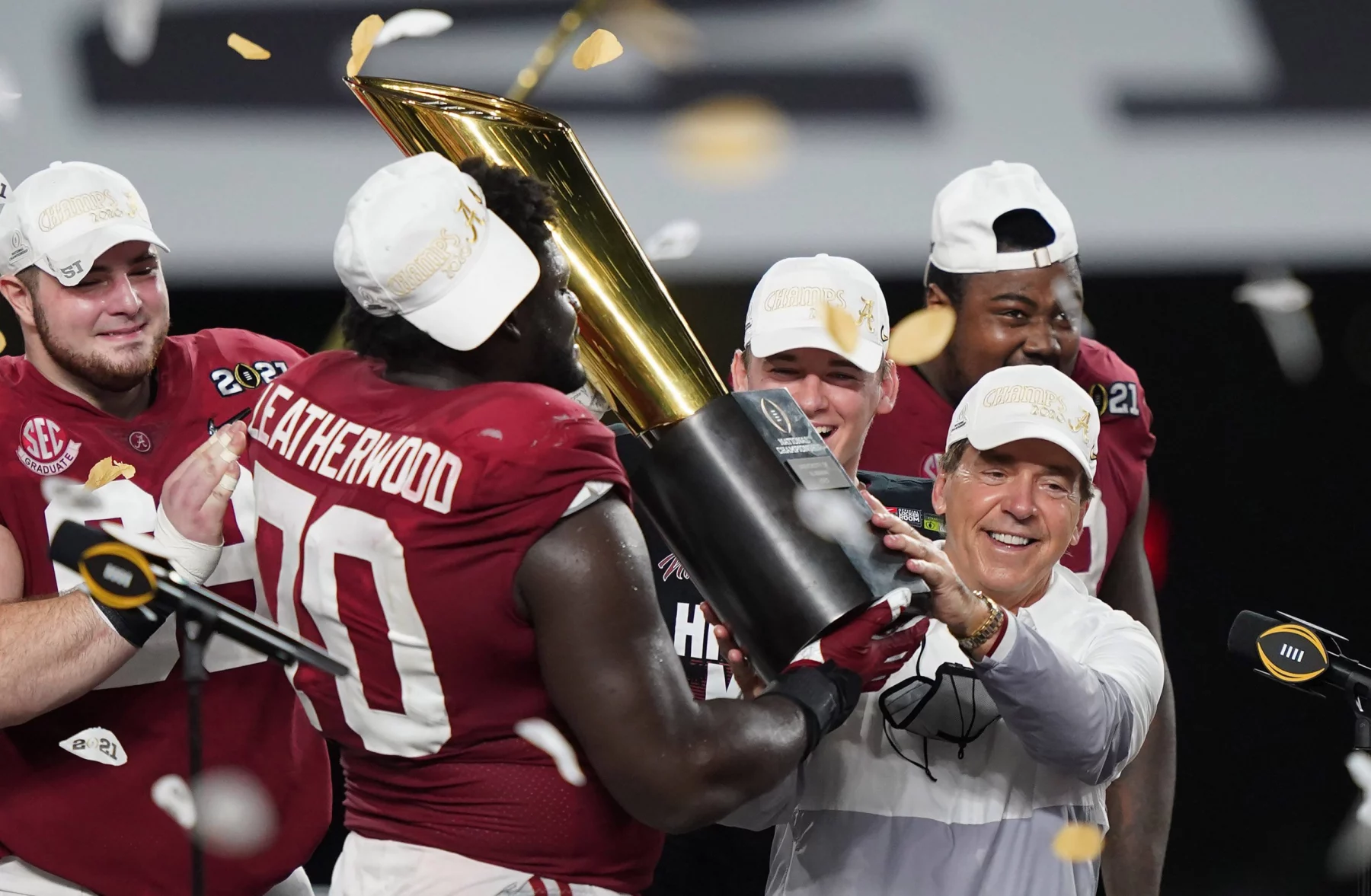 Kalen DeBoer will mit Alabama zurück an die Spitze. Eine Footballmannschaft in roten Trikots feiert den Gewinn der Nationalmeisterschaft. Die Spieler tragen weiße Hüte und halten einen großen goldenen Pokal in den Händen, während Konfetti um sie herum regnet. Ein Mann in weißer Jacke hilft, den Pokal zu halten, und lächelt zwischen den Spielern auf einer Bühne mit schwarz-weißem Hintergrund. Diese Beschreibung wurde mit der FootballR KI automatisch generiert.