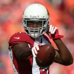 Patrick Peterson in der Uniform der Arizona Cardinals, mit weißem Helm und roten Handschuhen, bereitet sich auf einen Pass vor. Er ist konzentriert, hat seinen rechten Arm nach hinten gestreckt und hält den Football fest umklammert. Im Hintergrund ist eine verschwommene Menschenmenge in leuchtenden Orangetönen zu sehen, die eine lebhafte Atmosphäre suggeriert. Diese Beschreibung wurde mit der FootballR KI automatisch generiert.