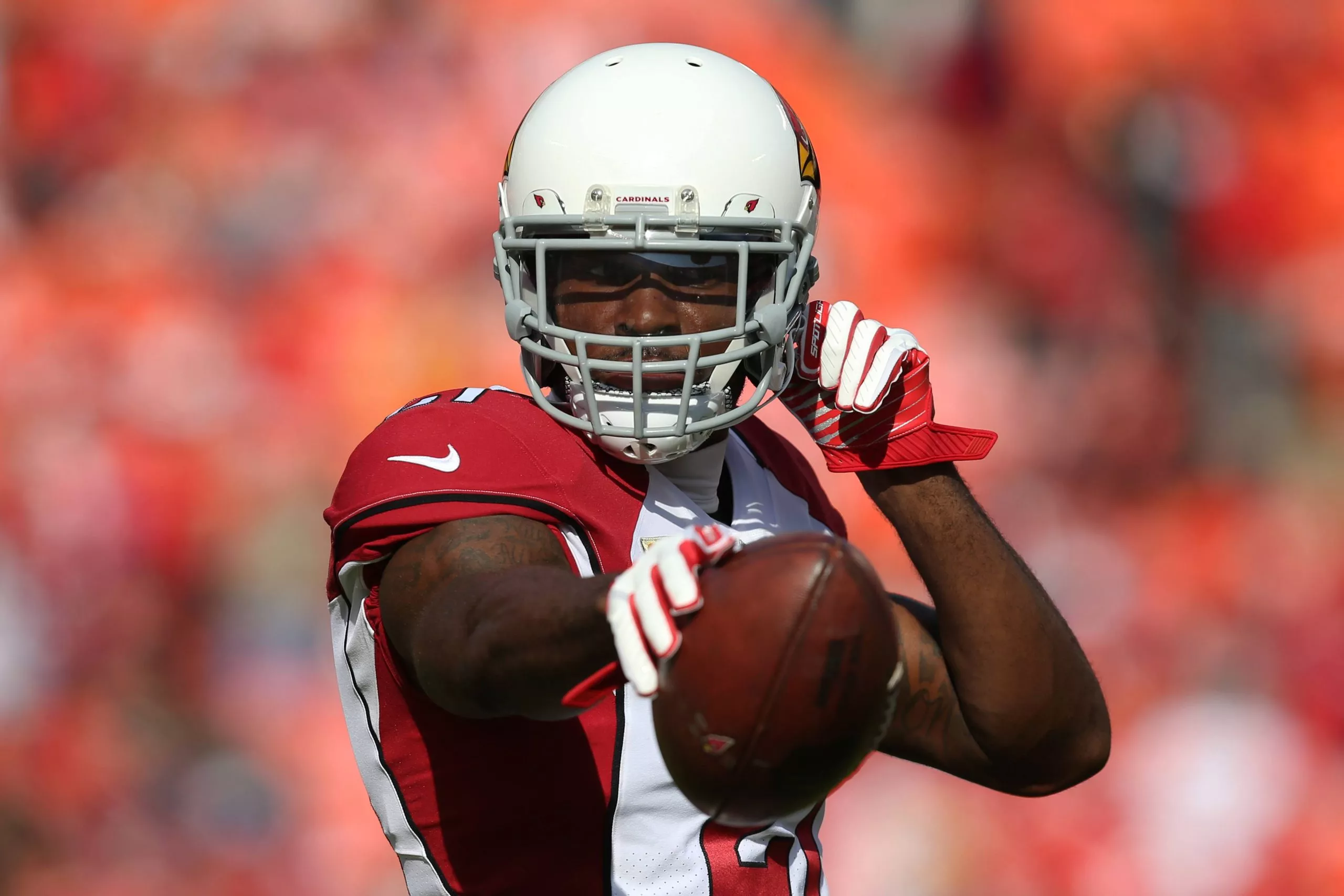Patrick Peterson in der Uniform der Arizona Cardinals, mit weißem Helm und roten Handschuhen, bereitet sich auf einen Pass vor. Er ist konzentriert, hat seinen rechten Arm nach hinten gestreckt und hält den Football fest umklammert. Im Hintergrund ist eine verschwommene Menschenmenge in leuchtenden Orangetönen zu sehen, die eine lebhafte Atmosphäre suggeriert. Diese Beschreibung wurde mit der FootballR KI automatisch generiert.
