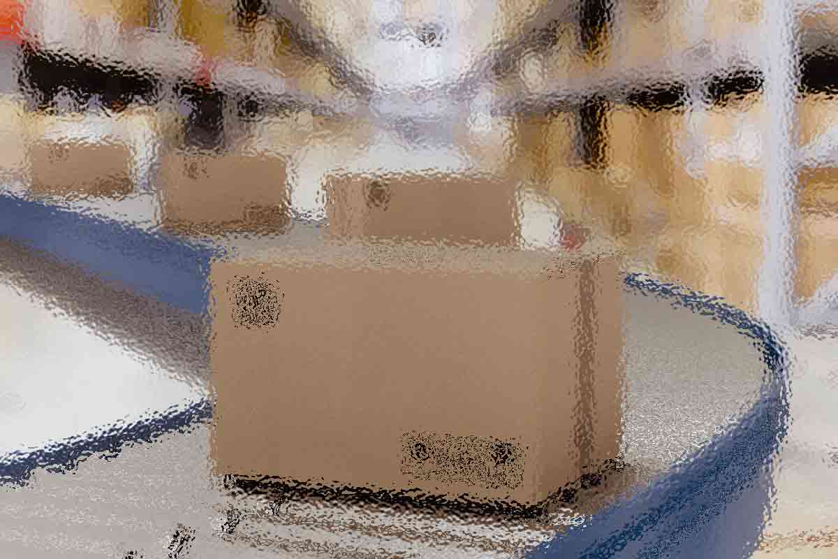 Ein Karton bewegt sich auf einem Förderband in einem Lagerhaus mit hohen Regalen, die mit ähnlichen Kartons gefüllt sind. Der Hintergrund erscheint unscharf, wodurch der Karton im Vordergrund hervorgehoben wird. Die Kulisse lässt auf ein geschäftiges Logistik- oder Vertriebszentrum schließen. Diese Beschreibung wurde mit der FootballR KI automatisch generiert.