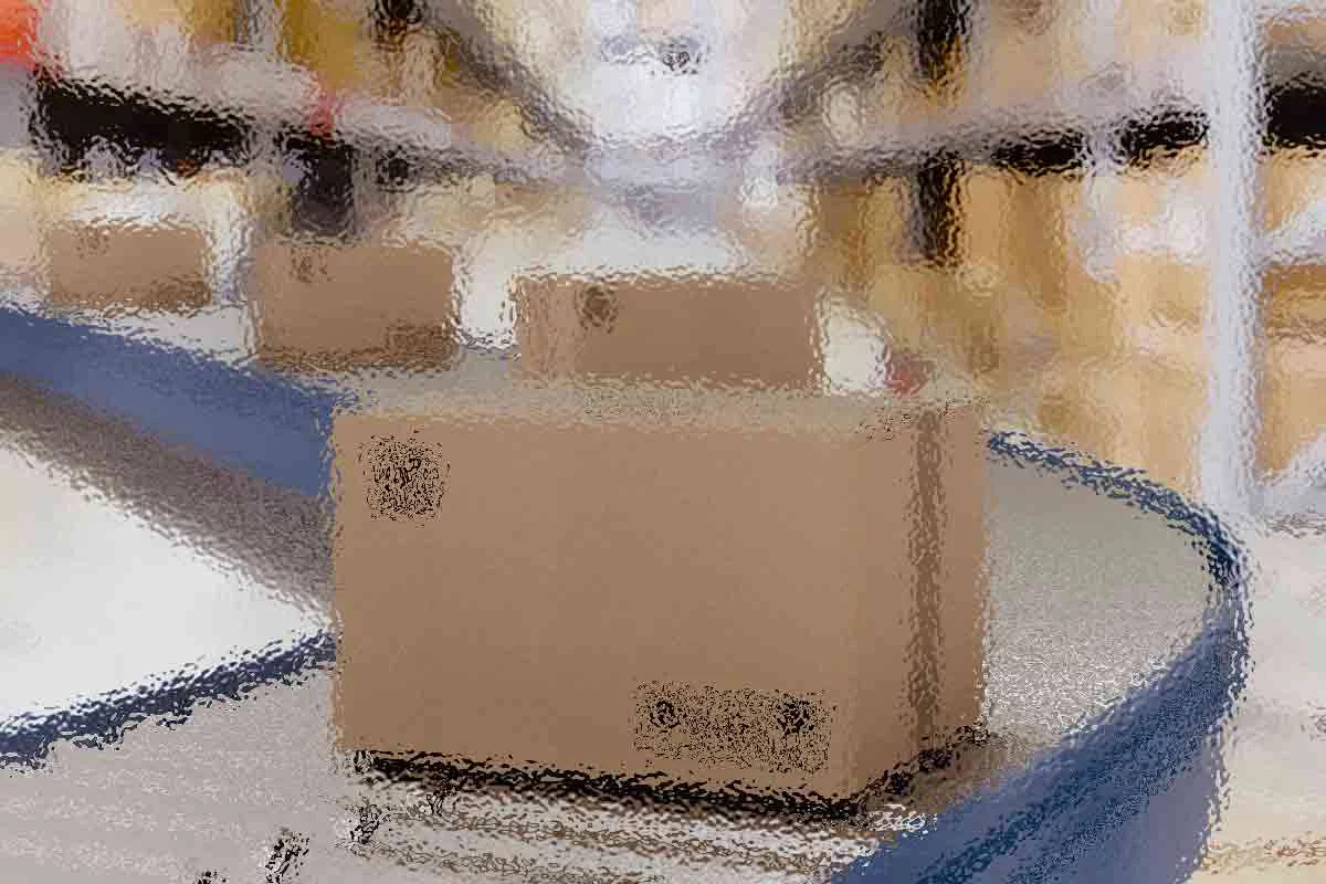 Ein Karton bewegt sich auf einem Förderband in einem Lagerhaus mit hohen Regalen, die mit ähnlichen Kartons gefüllt sind. Der Hintergrund erscheint unscharf, wodurch der Karton im Vordergrund hervorgehoben wird. Die Kulisse lässt auf ein geschäftiges Logistik- oder Vertriebszentrum schließen. Diese Beschreibung wurde mit der FootballR KI automatisch generiert.