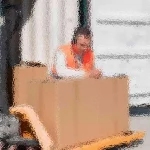 Unscharfes Bild einer Person mit orangefarbener Sicherheitsweste, die in einem großen Karton auf einer Palette sitzt. Der Karton steht auf einem orangefarbenen Palettenwagen vor einem Versandcontainer, was an eine Lagerhalle oder eine Laderampe erinnert. Diese Beschreibung wurde mit der FootballR KI automatisch generiert.