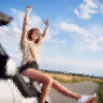 Eine Person sitzt auf einem geparkten Auto mit offenem Verdeck und streckt vor einem Hintergrund aus blauem Himmel und vereinzelten Wolken freudig die Arme aus. Sie trägt ein helles Hemd und Shorts. Die Szene vermittelt Freiheit und Glück an einem hellen, sonnigen Tag. Diese Beschreibung wurde mit der FootballR KI automatisch generiert.