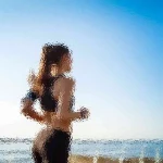 Eine Person mit langen Haaren joggt am Strand entlang. Sie trägt Sportkleidung und die Szene ist so gefiltert, dass sie abstrakt und verschwommen erscheint. Der Himmel ist klar und blau und im Hintergrund ist das Meer unter einer hellen Sonne teilweise sichtbar. Diese Beschreibung wurde mit der FootballR KI automatisch generiert.