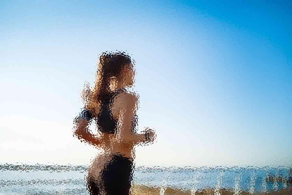 Eine Person mit langen Haaren joggt am Strand entlang. Sie trägt Sportkleidung und die Szene ist so gefiltert, dass sie abstrakt und verschwommen erscheint. Der Himmel ist klar und blau und im Hintergrund ist das Meer unter einer hellen Sonne teilweise sichtbar. Diese Beschreibung wurde mit der FootballR KI automatisch generiert.