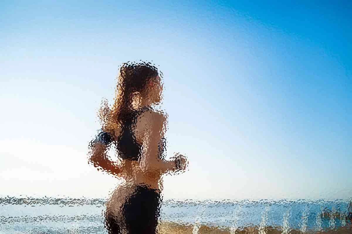 Eine Person mit langen Haaren joggt am Strand entlang. Sie trägt Sportkleidung und die Szene ist so gefiltert, dass sie abstrakt und verschwommen erscheint. Der Himmel ist klar und blau und im Hintergrund ist das Meer unter einer hellen Sonne teilweise sichtbar. Diese Beschreibung wurde mit der FootballR KI automatisch generiert.