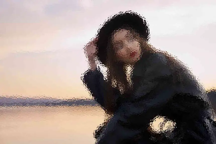 Ein verschwommenes künstlerisches Bild einer Person mit schwarzem Hut und Mantel, die bei Sonnenuntergang an einem ruhigen Gewässer sitzt. Der Himmel ist sanft mit Orange- und Lilatönen beleuchtet, die sich im Wasser spiegeln. Die Person scheint in die Kamera zu blicken, was eine heitere und besinnliche Atmosphäre schafft. Diese Beschreibung wurde mit der FootballR KI automatisch generiert.