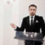 Ein Mann im Anzug steht hinter einem Podium und gestikuliert mit seiner rechten Hand. Das Bild ist stark verschwommen, sodass Details und Gesichtszüge nicht zu erkennen sind. Der Hintergrund ist eine schlichte, helle Wand. Diese Beschreibung wurde mit der FootballR KI automatisch generiert.