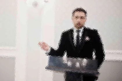Ein Mann im Anzug steht hinter einem Podium und gestikuliert mit seiner rechten Hand. Das Bild ist stark verschwommen, sodass Details und Gesichtszüge nicht zu erkennen sind. Der Hintergrund ist eine schlichte, helle Wand. Diese Beschreibung wurde mit der FootballR KI automatisch generiert.