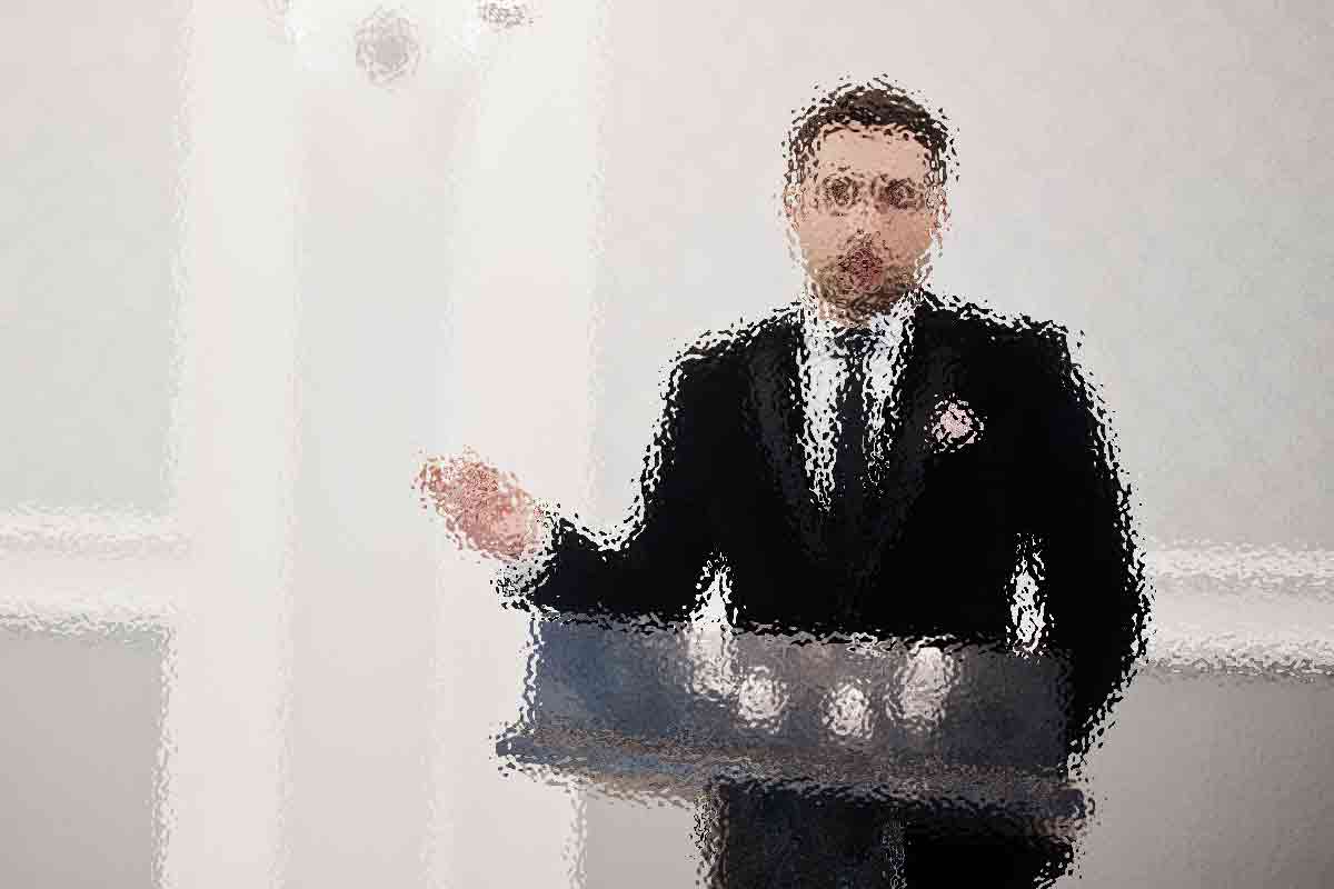 Ein Mann im Anzug steht hinter einem Podium und gestikuliert mit seiner rechten Hand. Das Bild ist stark verschwommen, sodass Details und Gesichtszüge nicht zu erkennen sind. Der Hintergrund ist eine schlichte, helle Wand. Diese Beschreibung wurde mit der FootballR KI automatisch generiert.