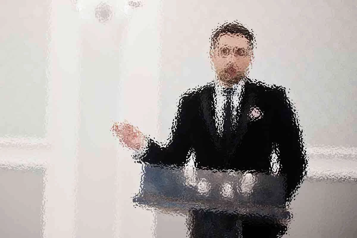 Ein Mann im Anzug steht hinter einem Podium und gestikuliert mit seiner rechten Hand. Das Bild ist stark verschwommen, sodass Details und Gesichtszüge nicht zu erkennen sind. Der Hintergrund ist eine schlichte, helle Wand. Diese Beschreibung wurde mit der FootballR KI automatisch generiert.