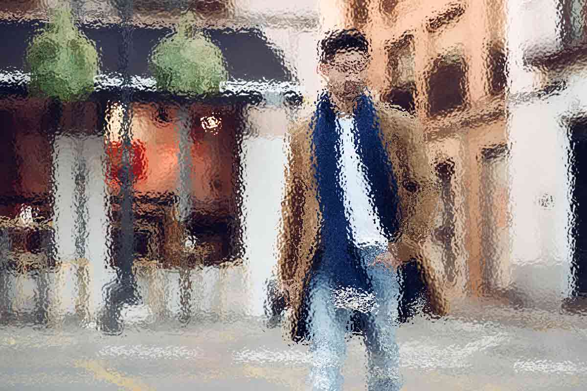 Eine Person in braunem Mantel, blauem Schal und Jeans geht auf einer Straße in einer verschwommenen Stadtlandschaft. Im Hintergrund sind Gebäude mit Ladenfronten zu sehen. Das Bild hat einen gemalten Effekt und verleiht der Szene eine weiche, künstlerische Note. Diese Beschreibung wurde mit der FootballR KI automatisch generiert.