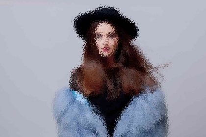 Eine Person mit langen Haaren, die einen schwarzen Hut und einen blauen, flauschigen Mantel trägt, steht vor einem hellgrauen Hintergrund. Das Bild ist absichtlich unscharf, wodurch ein impressionistischer Effekt entsteht, detaillierte Gesichtszüge verdeckt und die Gesamtform und -farben hervorgehoben werden. Diese Beschreibung wurde mit der FootballR KI automatisch generiert.