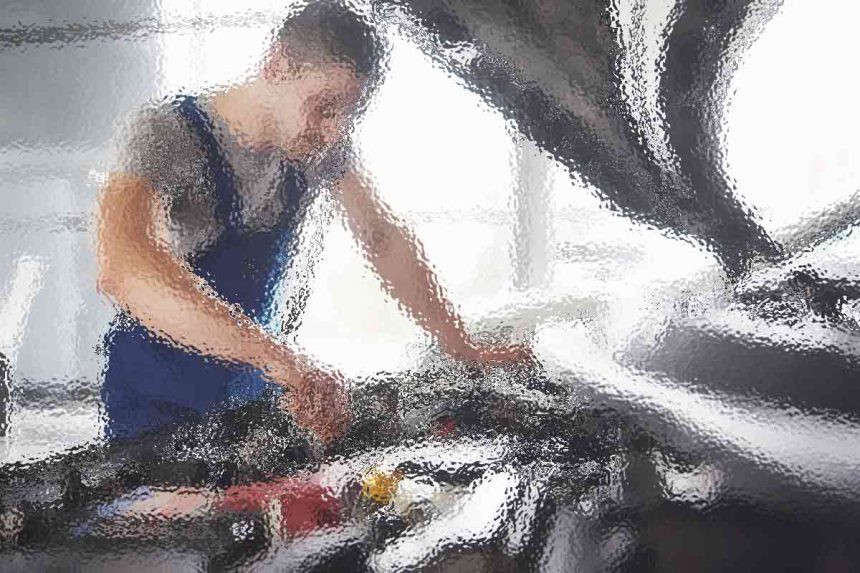 Ein Mechaniker im blauen Overall beugt sich über eine offene Motorhaube und arbeitet am Motor. Der Hintergrund ist unscharf und suggeriert eine Garagenumgebung mit natürlichem Licht, das durch die Fenster einfällt. Der Fokus liegt auf der aufmerksamen Haltung und den Händen des Mechanikers, wodurch die Konzentration auf das Fahrzeug betont wird. Diese Beschreibung wurde mit der FootballR KI automatisch generiert.