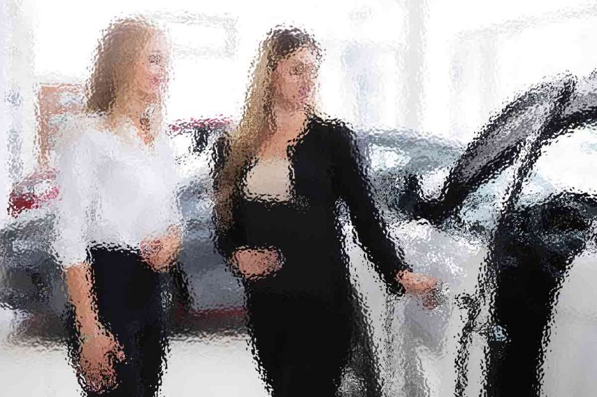 Zwei Frauen stehen neben einem Auto in einem Ausstellungsraum. Eine Frau, in schwarzem Outfit, öffnet die Autotür. Die andere Frau, in weißem Hemd und dunkler Hose, beobachtet das Auto. Im hell erleuchteten Hintergrund sind mehrere andere Fahrzeuge zu sehen. Diese Beschreibung wurde mit der FootballR KI automatisch generiert.