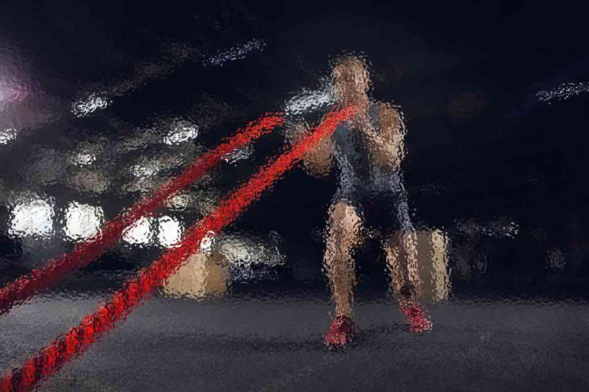 Eine Person in Sportkleidung trainiert in einem Fitnessstudio mit Battle Ropes. Die Person hockt und bewegt die roten Seile energisch in einem schwach beleuchteten Raum, der von sanften Deckenlampen erhellt wird. Diese Beschreibung wurde mit der FootballR KI automatisch generiert.