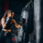 Eine Frau mit langen Haaren, schwarzen Boxhandschuhen und einem schwarzen Outfit, schlägt in einem schwach beleuchteten Fitnessstudio konzentriert auf einen großen schwarzen Boxsack ein. Das Bild hat einen verschwommenen, gemalten Effekt, der ihrer Trainingseinheit ein Gefühl von Bewegung und Energie verleiht. Diese Beschreibung wurde mit der FootballR KI automatisch generiert.