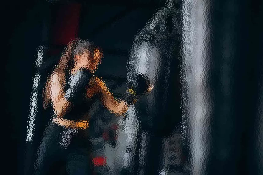 Eine Frau mit langen Haaren, schwarzen Boxhandschuhen und einem schwarzen Outfit, schlägt in einem schwach beleuchteten Fitnessstudio konzentriert auf einen großen schwarzen Boxsack ein. Das Bild hat einen verschwommenen, gemalten Effekt, der ihrer Trainingseinheit ein Gefühl von Bewegung und Energie verleiht. Diese Beschreibung wurde mit der FootballR KI automatisch generiert.