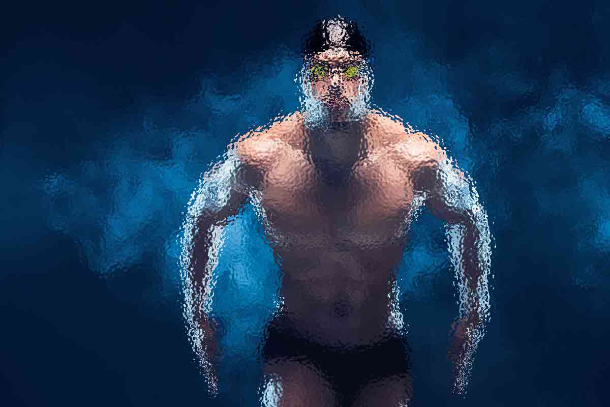 Ein Schwimmer mit Schwimmbrille, Badekappe und Badehose wird unter Wasser aufgenommen. Das Bild ist mit einem verschwommenen, künstlerischen Effekt stilisiert, der den muskulösen Körperbau und die dynamische Bewegung vor einem tiefblauen Hintergrund betont. Blasen und Lichtreflexionen verstärken die Unterwasseratmosphäre. Diese Beschreibung wurde mit der FootballR KI automatisch generiert.