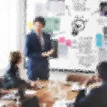 Ein verschwommenes Bild eines Leiters bei einem Geschäftstreffen. Die Person steht neben einer großen Tafel voller bunter Notizen und Diagramme und präsentiert vier sitzenden Kollegen etwas. Der Raum ist gut beleuchtet, da natürliches Licht durch ein großes Fenster fällt. Diese Beschreibung wurde mit der FootballR KI automatisch generiert.