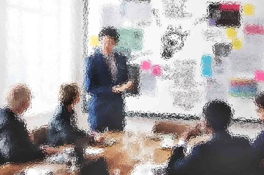 Ein verschwommenes Bild eines Leiters bei einem Geschäftstreffen. Die Person steht neben einer großen Tafel voller bunter Notizen und Diagramme und präsentiert vier sitzenden Kollegen etwas. Der Raum ist gut beleuchtet, da natürliches Licht durch ein großes Fenster fällt. Diese Beschreibung wurde mit der FootballR KI automatisch generiert.