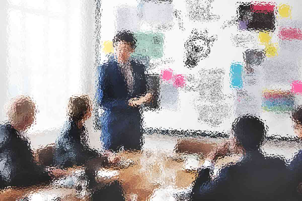 Ein verschwommenes Bild eines Leiters bei einem Geschäftstreffen. Die Person steht neben einer großen Tafel voller bunter Notizen und Diagramme und präsentiert vier sitzenden Kollegen etwas. Der Raum ist gut beleuchtet, da natürliches Licht durch ein großes Fenster fällt. Diese Beschreibung wurde mit der FootballR KI automatisch generiert.