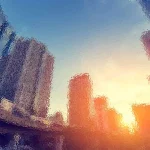 Abstrakte Stadtlandschaft bei Sonnenuntergang mit verschwommenen, verzerrten Wolkenkratzern. Die Sonne geht am Horizont unter und wirft einen orangefarbenen Schimmer auf die Gebäude. Der Himmel wechselt von Blau zu einem warmen Gelb und Orange und schafft so eine verträumte, surreale Atmosphäre. Diese Beschreibung wurde mit der FootballR KI automatisch generiert.