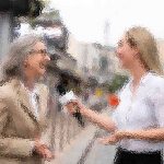 Zwei Frauen in einer unscharfen Szene im Freien interagieren miteinander. Eine hält ein Mikrofon und scheint die andere zu interviewen, die eine Brille und einen beigen Blazer trägt. Beide Frauen lächeln und unterhalten sich. Sie stehen auf einer Straße mit Gebäuden im Hintergrund. Diese Beschreibung wurde mit der FootballR KI automatisch generiert.