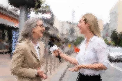 Zwei Frauen in einer unscharfen Szene im Freien interagieren miteinander. Eine hält ein Mikrofon und scheint die andere zu interviewen, die eine Brille und einen beigen Blazer trägt. Beide Frauen lächeln und unterhalten sich. Sie stehen auf einer Straße mit Gebäuden im Hintergrund. Diese Beschreibung wurde mit der FootballR KI automatisch generiert.