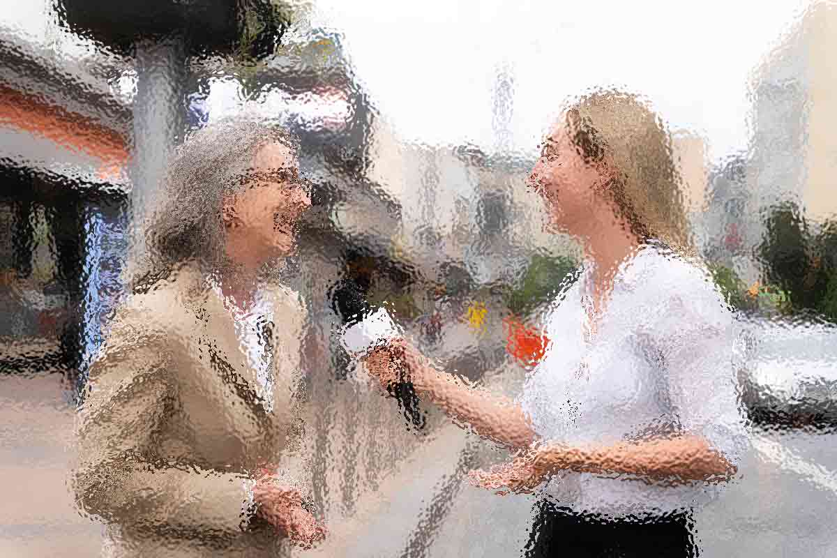 Zwei Frauen in einer unscharfen Szene im Freien interagieren miteinander. Eine hält ein Mikrofon und scheint die andere zu interviewen, die eine Brille und einen beigen Blazer trägt. Beide Frauen lächeln und unterhalten sich. Sie stehen auf einer Straße mit Gebäuden im Hintergrund. Diese Beschreibung wurde mit der FootballR KI automatisch generiert.