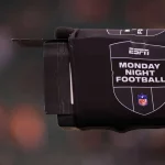ESPN kauft NFL Network - NFL-Quoten - Nahaufnahme einer Fernsehkamera, die mit einer schwarzen Hülle bedeckt ist, auf der das ESPN-Logo und der Text "Monday Night Football" zusammen mit dem NFL-Schild abgebildet sind, um die Rechte von ESPN zur Übertragung des Monday Night Wild Card Game hervorzuheben; unscharfer Stadionhintergrund. Diese Beschreibung wurde mit der FootballR KI automatisch generiert.