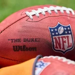 Franchise Tag - NFL Woche 12: Nahaufnahme eines offiziellen NFL-Footballs auf einem Rasenfeld. Der Ball zeigt das NFL-Logo und die Wörter „THE DUKE“ und „Wilson“. Er hat weiße Schnürsenkel und eine strukturierte braune Oberfläche. Im Vordergrund ist ein Teil eines anderen Footballs teilweise sichtbar. Diese Beschreibung wurde mit der FootballR KI automatisch generiert.