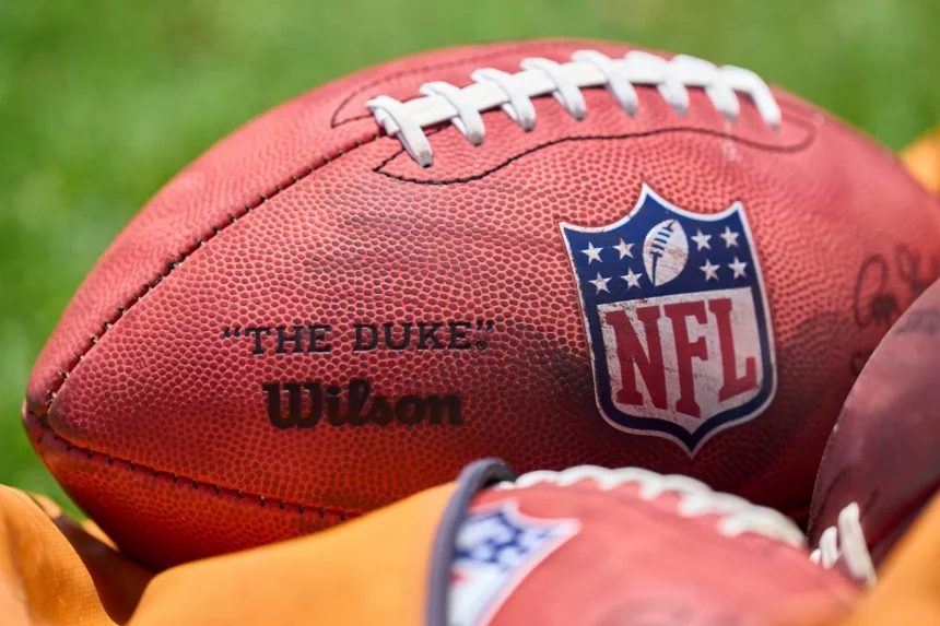 Franchise Tag - NFL Woche 12: Nahaufnahme eines offiziellen NFL-Footballs auf einem Rasenfeld. Der Ball zeigt das NFL-Logo und die Wörter „THE DUKE“ und „Wilson“. Er hat weiße Schnürsenkel und eine strukturierte braune Oberfläche. Im Vordergrund ist ein Teil eines anderen Footballs teilweise sichtbar. Diese Beschreibung wurde mit der FootballR KI automatisch generiert.