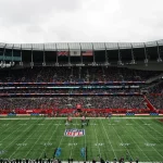 American Football - International Series - In einem voll besetzten Stadion findet ein NFL-Spiel statt. Auf Bildschirmen sind zwei große US-Flaggen zu sehen, darüber wehen die Flaggen Großbritanniens und der USA. Die Spieler stehen auf dem Spielfeld in Reih und Glied, während Fans die Tribünen unter einem bewölkten Himmel füllen und so die internationale Atmosphäre unterstreichen. Diese Beschreibung wurde mit der FootballR KI automatisch generiert.