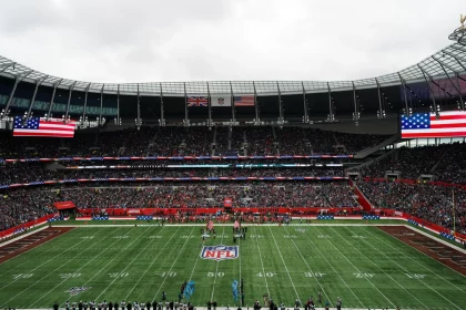 American Football - International Series - In einem voll besetzten Stadion findet ein NFL-Spiel statt. Auf Bildschirmen sind zwei große US-Flaggen zu sehen, darüber wehen die Flaggen Großbritanniens und der USA. Die Spieler stehen auf dem Spielfeld in Reih und Glied, während Fans die Tribünen unter einem bewölkten Himmel füllen und so die internationale Atmosphäre unterstreichen. Diese Beschreibung wurde mit der FootballR KI automatisch generiert.