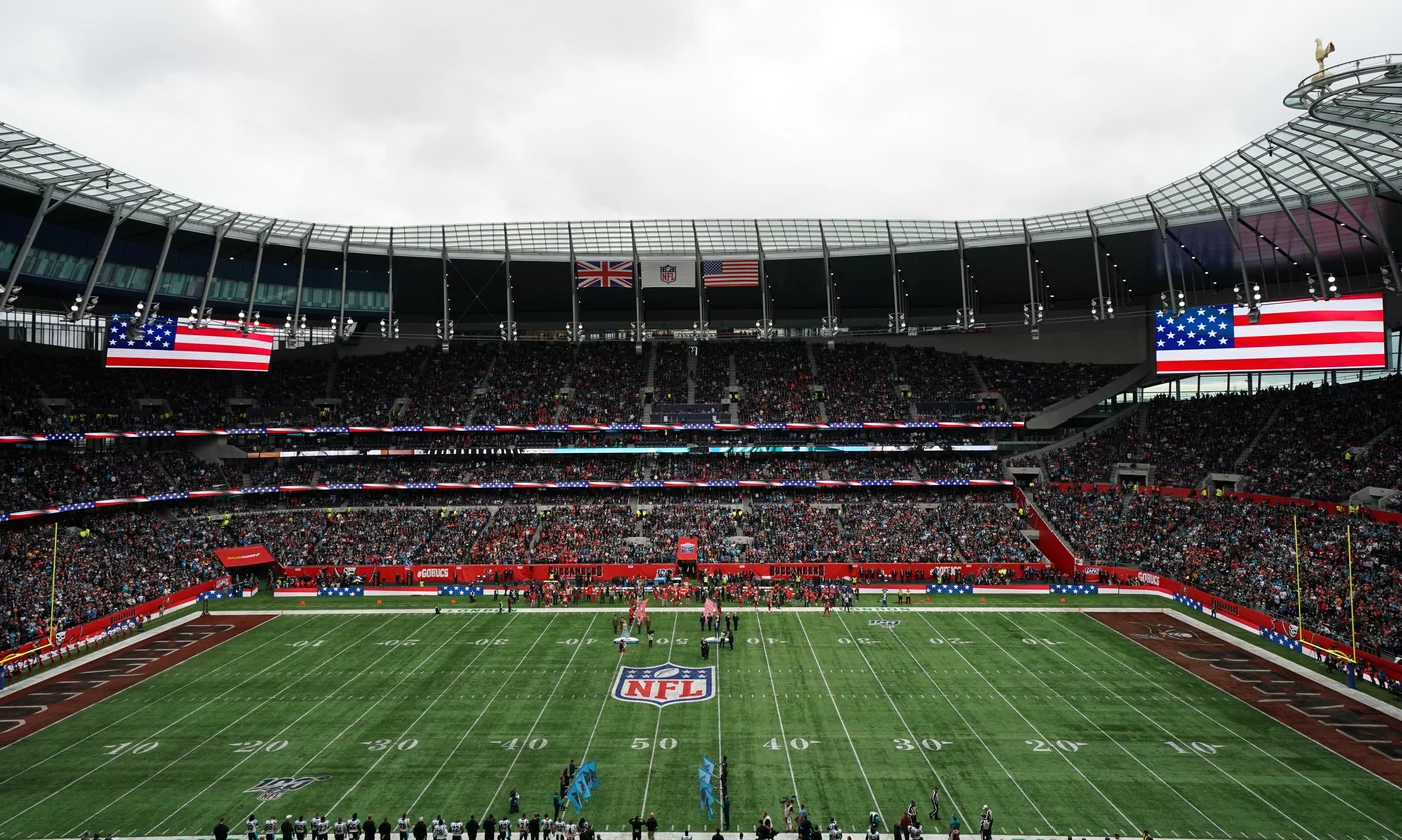 American Football - International Series - In einem voll besetzten Stadion findet ein NFL-Spiel statt. Auf Bildschirmen sind zwei große US-Flaggen zu sehen, darüber wehen die Flaggen Großbritanniens und der USA. Die Spieler stehen auf dem Spielfeld in Reih und Glied, während Fans die Tribünen unter einem bewölkten Himmel füllen und so die internationale Atmosphäre unterstreichen. Diese Beschreibung wurde mit der FootballR KI automatisch generiert.
