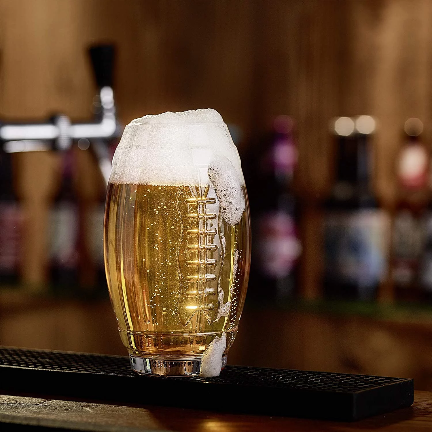 FootballR - NFL - Ein klares, schaumiges Bier wird in ein footballförmiges Glas mit strukturiertem Design gegossen, das auf einer Barmatte steht. Im Hintergrund sind unscharfe Zapfhähne und Flaschen zu sehen, was an eine Bar oder Kneipe erinnert. Man sieht Blasen aufsteigen und Schaum läuft leicht über den Rand des Glases. Diese Beschreibung wurde automatisch generiert.