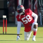 Shaquil Barrett auch Shaq Barrett genannt in einem roten Trikot mit der Nummer 58 und weißen Hosen kniet beim Training auf dem Rasen. Tochter Barretts typischer Stil wird durch seinen Helm mit dunklem Visier deutlich. Im Hintergrund sind Ausrüstung und Sitzgelegenheiten zu sehen. Diese Beschreibung wurde mit der FootballR KI automatisch generiert.