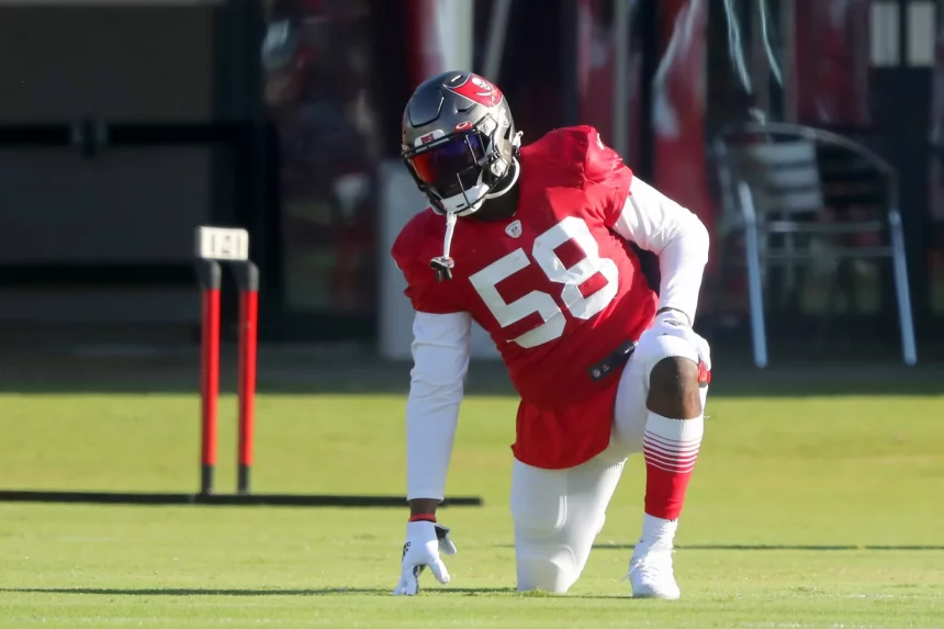 Shaquil Barrett auch Shaq Barrett genannt in einem roten Trikot mit der Nummer 58 und weißen Hosen kniet beim Training auf dem Rasen. Tochter Barretts typischer Stil wird durch seinen Helm mit dunklem Visier deutlich. Im Hintergrund sind Ausrüstung und Sitzgelegenheiten zu sehen. Diese Beschreibung wurde mit der FootballR KI automatisch generiert.