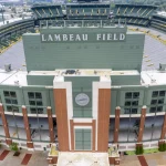 FootballR - NFL - Luftaufnahme des Lambeau Field in Green Bay, einem Freiluftstadion mit grüner Fassade, Ziegelakzenten und zahlreichen Sitzbereichen. Auf jeder Seite des Gebäudes befindet sich eine amerikanische Flagge. Über den Türen des Haupteingangs befindet sich eine große Uhr. Der Austragungsort des NFL Draft 2025 ist von Bäumen und Gehwegen umgeben. Diese Beschreibung wurde automatisch generiert.