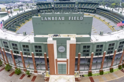 FootballR - NFL - Luftaufnahme des Lambeau Field in Green Bay, einem Freiluftstadion mit grüner Fassade, Ziegelakzenten und zahlreichen Sitzbereichen. Auf jeder Seite des Gebäudes befindet sich eine amerikanische Flagge. Über den Türen des Haupteingangs befindet sich eine große Uhr. Der Austragungsort des NFL Draft 2025 ist von Bäumen und Gehwegen umgeben. Diese Beschreibung wurde automatisch generiert.
