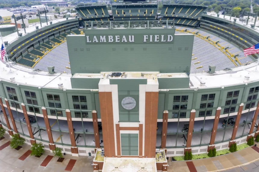 FootballR - NFL - Luftaufnahme des Lambeau Field in Green Bay, einem Freiluftstadion mit grüner Fassade, Ziegelakzenten und zahlreichen Sitzbereichen. Auf jeder Seite des Gebäudes befindet sich eine amerikanische Flagge. Über den Türen des Haupteingangs befindet sich eine große Uhr. Der Austragungsort des NFL Draft 2025 ist von Bäumen und Gehwegen umgeben. Diese Beschreibung wurde automatisch generiert.