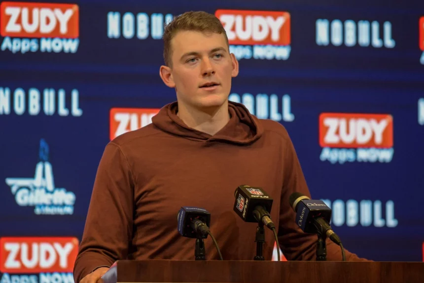 FootballR - NFL - New England Patriots QB Mac Jones steht bei einer Pressekonferenz vor einem Mikrofon.