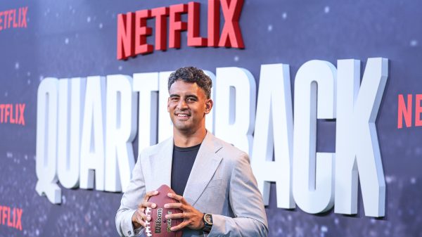 FootballR - NFL - Ein Mann im Anzug hält einen Football in der Hand, während er vor einem Netflix-Schild posiert.